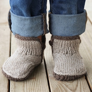 May include: A pair of knitted beige slippers with brown trim. The slippers are worn by a person wearing blue jeans.