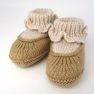 Op de afbeelding: Een paar gebreide babyschoentjes met een bruine zool en een beige gerimpelde bovenkant.