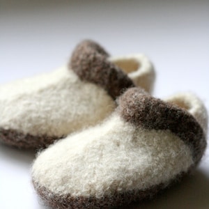 May include: A pair of white and brown knitted baby slippers. The slippers have a brown trim around the top and bottom.