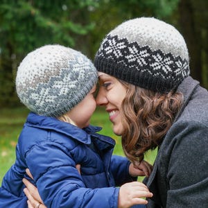 May include: A mother and child wearing matching grey and black knitted hats with a snowflake pattern. The hats are made of wool and have a soft, cosy feel.