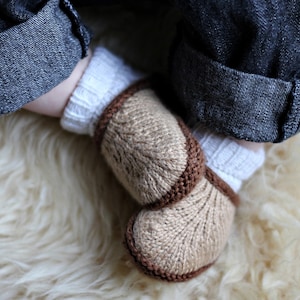 May include: A pair of knitted baby booties with brown trim. The booties are beige and have a textured knit pattern. The booties are lying on a white fluffy surface.