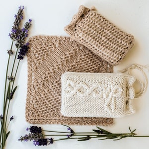 May include: Three knitted soap sacks, two in a light brown color and one in a white color. The white sack has the word "soap" knitted into it. The sacks are arranged on a white surface with lavender sprigs.