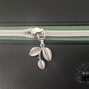 May include: A close-up of a black zipper with a silver zipper pull. The zipper pull is shaped like a small branch with three leaves.