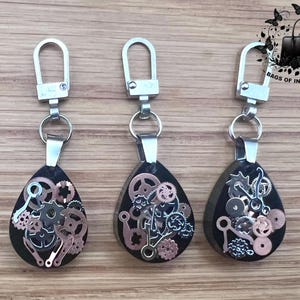 May include: Three teardrop-shaped keychains with silver-tone hardware. Each keychain features a black background with embedded gears and cogs in silver and copper tones. The keychains are displayed on a wooden surface.