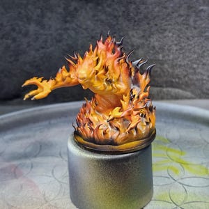 May include: A miniature figurine of a fiery elemental, with flames in shades of orange, yellow, and black. The figure is posed with one arm extended, and is mounted on a cylindrical base with a gradient finish.