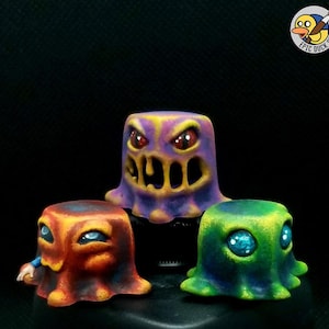 May include: Three colorful, stylized monster figurines. The purple monster has a wide, toothy grin and angry red eyes. The orange monster has blue eyes and a small arm. The green monster has large, sparkling blue eyes.