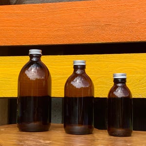 May include: Three amber glass bottles of varying sizes arranged in a row, each featuring a rounded body, a narrow neck, and a silver-coloured screw-top cap. The bottles are transparent, showing a dark brown tint, and have a smooth, cylindrical shape.