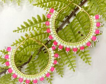 Pode incluir: Um par de brincos de argola dourados com contas rosa e brancas. Os brincos são adornados com contas em um fio verde.