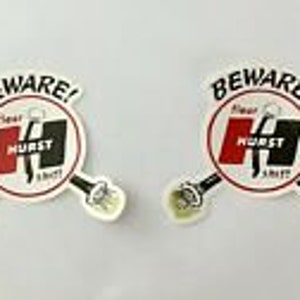 Peut inclure: Deux autocollants ronds blancs avec un logo Hurst rouge et noir et le texte "Beware! Hurst Shifters" en noir. Le logo présente un "H" stylisé avec un fond rouge et un contour noir.