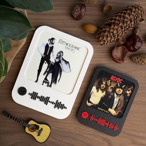 Op de afbeelding: Twee vierkante onderzetters met albumhoezen. Eén witte met Fleetwood Mac's 'Rumours' en één zwarte met AC/DC's 'Highway to Hell'. Beide hebben een Spotify-logo en een golfpatroon. Een kleine gele gitaar is ook aanwezig.