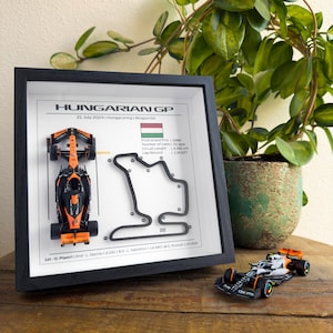 May include: A framed print of the Hungaroring race track with a miniature orange and black Formula 1 race car. The print includes the date of the race, the number of laps, the circuit length, and the lap record. The text "1st - O. Piastri 2nd - L. Norris +2.141 3rd - L. Hamilton +4.880 4th - G. Russell +20.805" is at the bottom of the print.