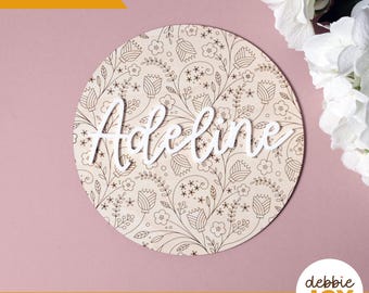 May include: A round wooden sign with the name "Adeline" in white script. The sign features a floral pattern and the text "Laser tested". The sign is on a pink surface.