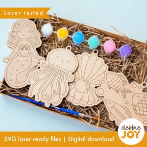 May include: A wooden craft kit featuring mermaid, jellyfish, seashell, and fish cutouts. The kit includes small paint pots in various colors and a paintbrush. The words "Laser tested" and "SVG laser ready files | Digital download" are visible.