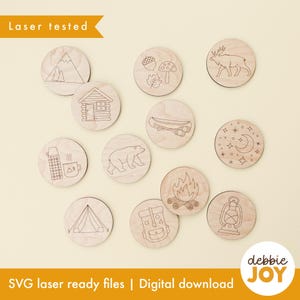 May include: A collection of eleven circular wooden discs featuring laser-engraved nature and camping designs, including a mountain, cabin, bear, canoe, campfire, lantern, backpack, tent, thermos, acorn, leaf, mushroom, moose, and moon with stars.