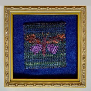 May include: A framed textile art piece featuring a butterfly design. The butterfly has purple, red, and orange details on a dark blue and green background. The artwork is set within a gold-colored frame and a blue mat.
