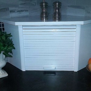 May include: White cabinet with a roll-up door, a white planter with green plants, and a bowl of fruit on a black countertop.