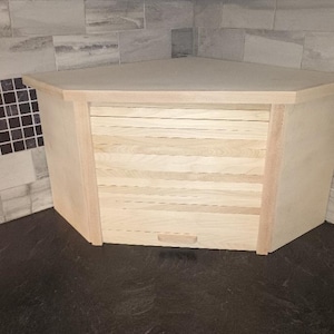 May include: A light-colored wooden corner bread box featuring a tambour door with horizontal slats and a small rectangular handle. The unit has a flat, triangular top surface and is designed to fit into a kitchen corner space.