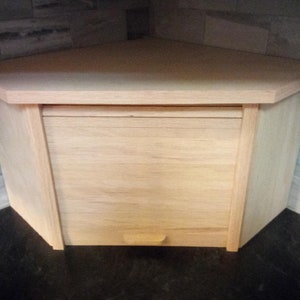 May include: A light wood corner bread box with a hinged door and a small handle. The bread box is sitting on a black countertop.