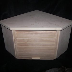 May include: A light-colored wooden bread box with a unique triangular shape. The box features a roll-top door and a small handle. The wood grain is visible, and the box appears to be unfinished.