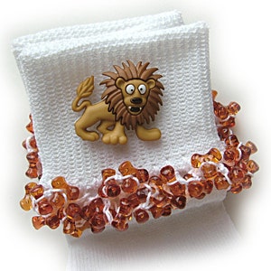 May include: A white knitted cuff featuring a brown plastic lion button with a cartoonish expression. The cuff is embellished with a border of small, translucent amber-colored beads shaped like bones, woven into the white fabric edge.