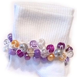May include: White ankle socks with a beaded trim. The trim is made of purple, pink, gold, and silver beads.