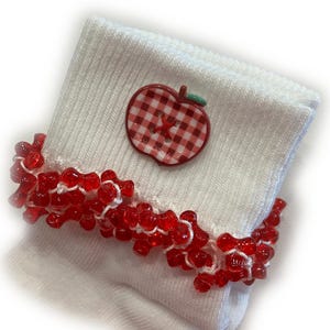 May include: White sock with a red beaded trim and a red gingham apple button. The sock is made of a ribbed knit material. The apple button has a red cross-stitch design.