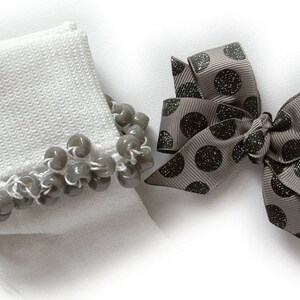 May include: A white sock featuring a cuff embellished with a row of glossy grey beads, paired with a grey hair bow made of ribbed fabric decorated with a pattern of dark, glittery polka dots. The items are presented against a plain white background.
