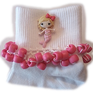 May include: White sock with a pink mermaid embellishment and a pink beaded trim. The mermaid has blonde hair and a pink bow. The trim is made of pink beads strung together with white thread.