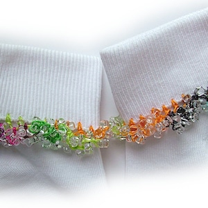 May include: Two white socks with colorful beaded trim. The trim features a variety of colors, including green, pink, orange, and black. The socks are made of a ribbed fabric.