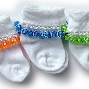 May include: Three pairs of white baby socks with colorful beaded trim. One pair has orange beads, another has blue beads, and the third has green beads. The socks are arranged on a white surface.