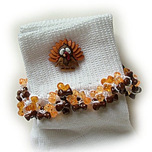 May include: White sock with a decorative beaded cuff in amber and brown. A small cartoon turkey button is attached. The sock has a textured, woven appearance. The cuff is decorated with amber and brown beads.