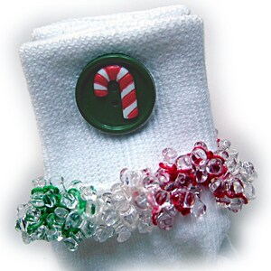 May include: White folded cloth napkin with a green button featuring a red and white candy cane design. A beaded napkin ring with clear, red, and green beads encircles the napkin, creating a festive holiday theme.