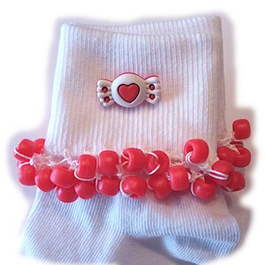May include: A pair of white ribbed ankle socks featuring a white and red candy-shaped charm with a pink heart center attached to the cuff. The cuff is decorated with a row of bright red plastic pony beads threaded onto white string.