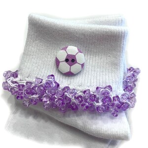 May include: A pair of white ribbed ankle socks featuring a decorative football button in white and purple. The cuff of each sock is embellished with a dense trim of translucent purple beads, creating a textured and layered appearance.