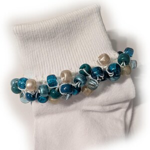 May include: A white sock with a beaded anklet. The anklet features a mix of blue and tan beads strung together with white thread. The beads are various shades of blue, from light to dark, and some are translucent.