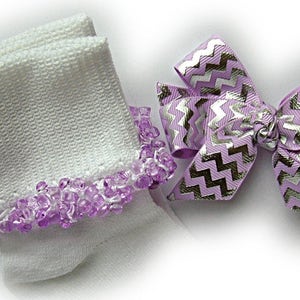 May include: A pair of white ankle socks featuring a cuff adorned with small, translucent purple beads, accompanied by a hair bow made of lavender ribbon with a metallic silver chevron pattern and a central knotted fabric detail.