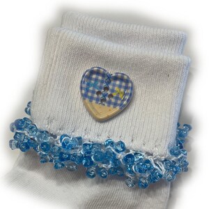 May include: A pair of white ribbed socks featuring a blue and white checkered heart-shaped button attached to the cuff. The cuff is decorated with a dense trim of small, translucent light blue beads that hang along the edge of the fabric.