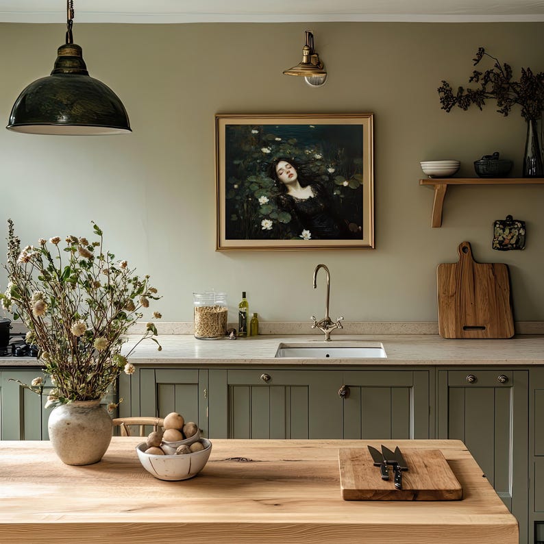 May include: A kitchen featuring olive green cabinets, a wooden worktop, and a framed painting. A large, dark green pendant light hangs above the wooden island. A vase of dried flowers and a bowl of fruit sit on the island.