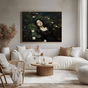May include: A framed art print shows a woman in a black dress amongst lily pads and white flowers. The artwork has a dark colour palette and is displayed above a white sofa in a living room setting.