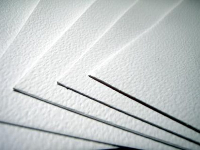 May include: Close-up of several sheets of white textured paper, fanned out. Three dark pencils are visible, with their tips pointing towards the upper right corner. The paper has a subtle, grainy texture.