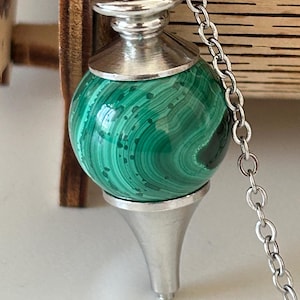 Peut inclure: Un pendule avec une sphère polie en malachite verte, suspendue par une chaîne en argent. Le pendule a des accents en métal argenté et une base conique. La malachite présente des motifs tourbillonnants naturels.