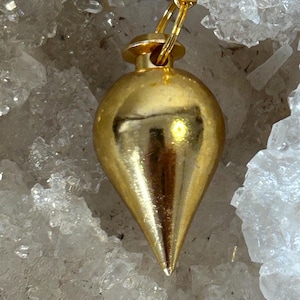 Op de afbeelding: Een goudkleurige pendel met een druppelvorm, opgehangen aan een gouden ketting. De pendel heeft een afgeronde bovenkant en een puntige onderkant. De achtergrond is een cluster van witte kristallen.