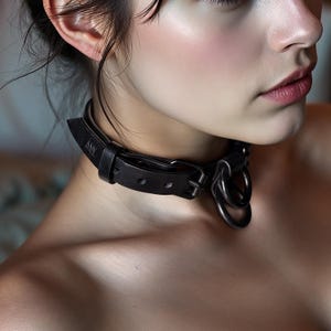 Può includere: Un choker in pelle nera con chiusura a fibbia e due anelli metallici. Il choker ha le lettere "AM" stampate sulla pelle. Il choker è indossato intorno al collo.