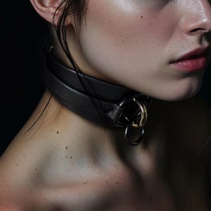 Könnte beinhalten: Ein schwarzer Lederkragen mit doppellagigem Design und Metallringen. Der Kragen wird um den Hals getragen, mit einem goldfarbenen Ring und einem größeren schwarzen Ring. Das Bild ist eine Nahaufnahme.
