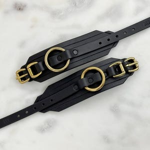 Könnte beinhalten: Zwei schwarze Leder-Handgelenkmanschetten mit goldfarbenen Metallbeschlägen. Jede Manschette hat einen großen goldenen Ring, eine Schnalle und einen Riemen mit mehreren Verstelllöchern. Die Manschetten werden auf einer weiß-grauen Marmoroberfläche präsentiert.