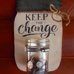 May include: A white wooden sign with the text "KEEP THE CHANGE" in black lettering. A glass jar with a metal clamp is attached to the sign and filled with coins.