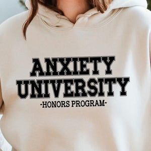 May include: A beige hoodie with the text "ANXIETY UNIVERSITY - HONORS PROGRAM" in black block letters. The hoodie has a front pocket and a hood. The person in the image is wearing a hat and sitting in a wicker chair.