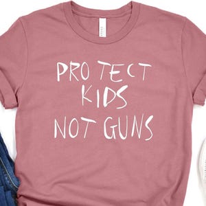 May include: A dusty rose t-shirt with the text "PROTECT KIDS NOT GUNS" in white. The shirt is paired with blue jeans and white slip-on shoes. The shirt is knotted at the bottom.