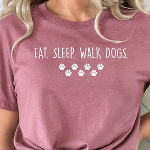 May include: A dusty rose t-shirt with the text "EAT. SLEEP. WALK DOGS." in white, along with paw print graphics. The shirt is paired with denim shorts. The model is standing in front of a neutral background.