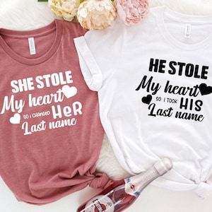 May include: Two t-shirts, one mauve and one white, with black text and heart graphics. The mauve shirt reads "She stole my heart so I changed her last name." The white shirt says "He stole my heart so I took his last name."